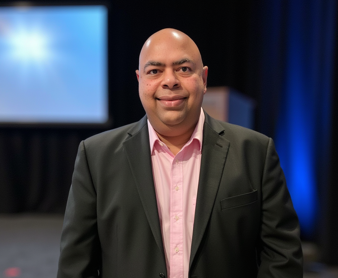 Ganesh Ariyur standing at a conference venue, in front of a presentation screen, ready to engage with the audience